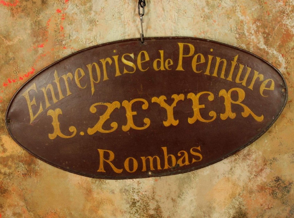 An oval dark brown metal trade sign featuring hand-painted yellow French lettering.