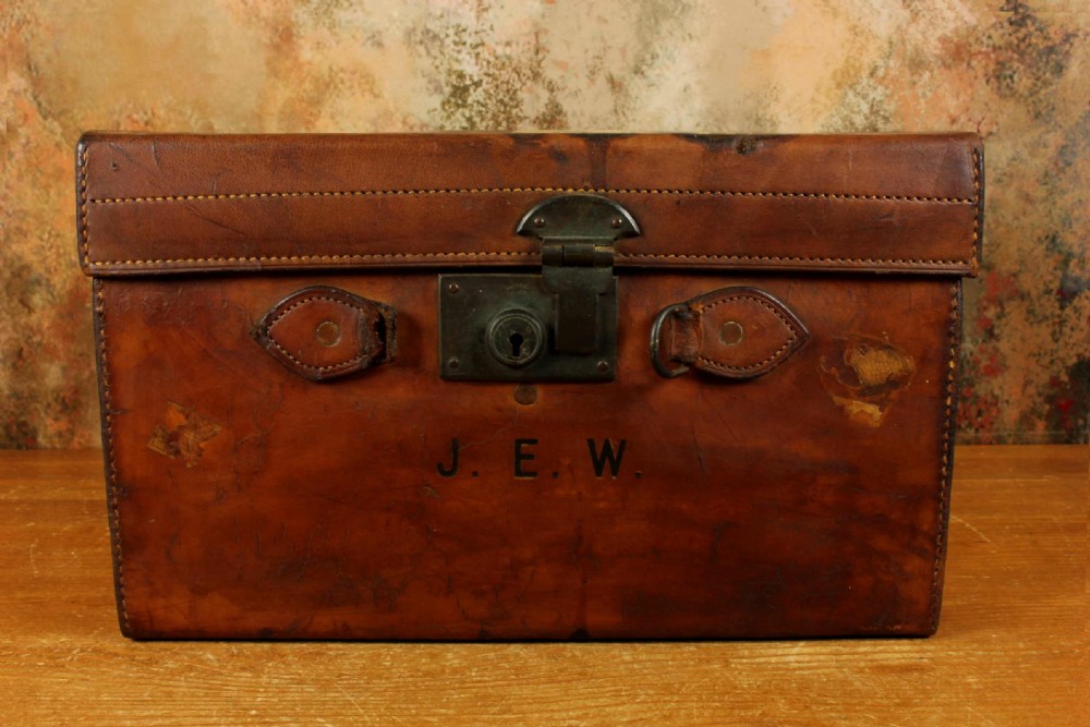 An antique tan leather hat box featuring an open lid, deep burgundy fabric lining with leather straps, a central brass lock, and initials J. E. W. stamped on the front. View III