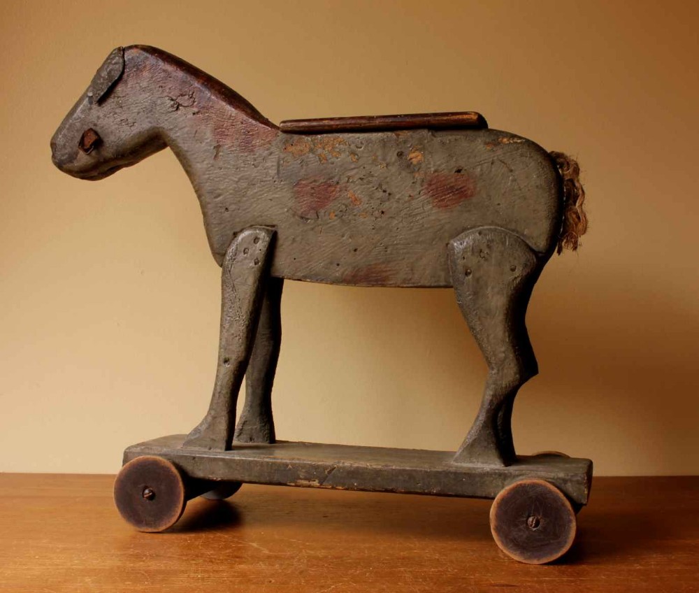 A carved wooden pull toy depicting a horse or donkey with distressed grey paint, mounted on a base with four small wooden wheels.