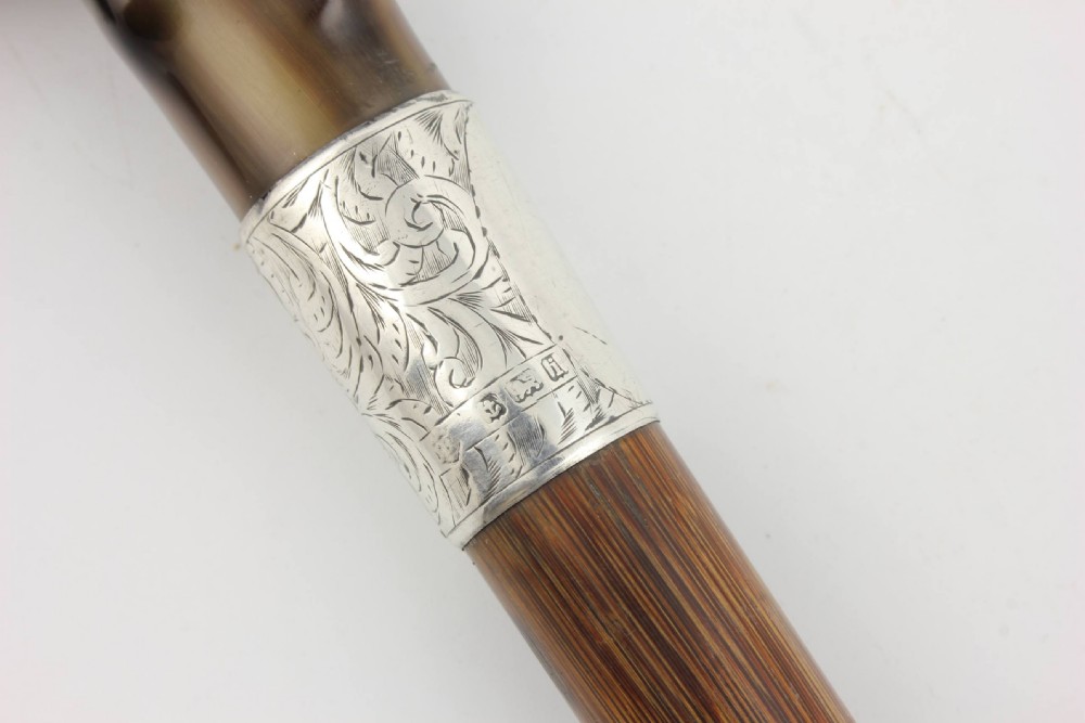 An Edwardian walking stick featuring a curved brown horn handle with a silver tip and an engraved sterling silver collar attached to a dark bamboo shaft. View V