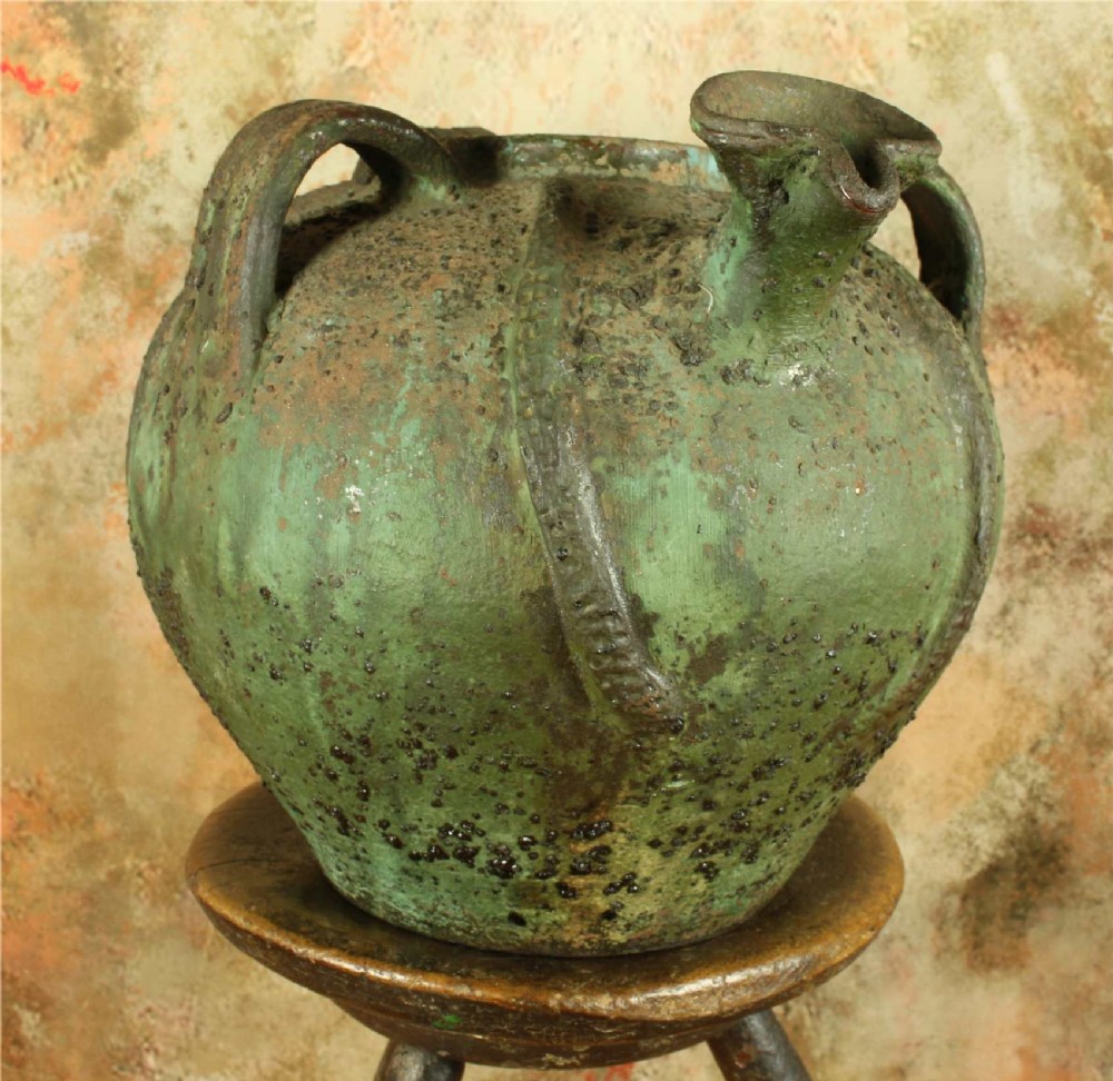 A bulbous antique green earthenware jug featuring three handles, a pour spout, and a heavily textured weathered glazed surface.