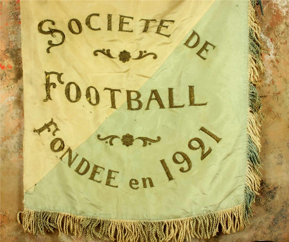 A square two-tone silk banner featuring pale green and cream diagonal panels, embroidered lettering reading Crossing FC Ganshoren, a central footballer figure, and fringe trim. View IV