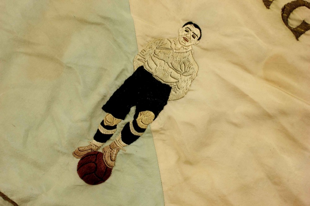 A square two-tone silk banner featuring pale green and cream diagonal panels, embroidered lettering reading Crossing FC Ganshoren, a central footballer figure, and fringe trim. View IX