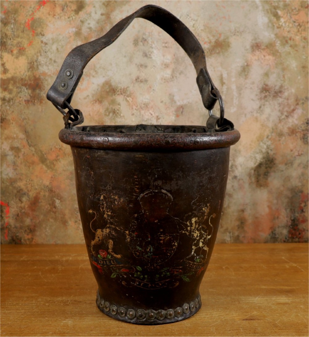 A dark brown Georgian leather fire bucket featuring a heavy strap handle, brass studs around the base, and a painted royal coat of arms with a lion and unicorn.