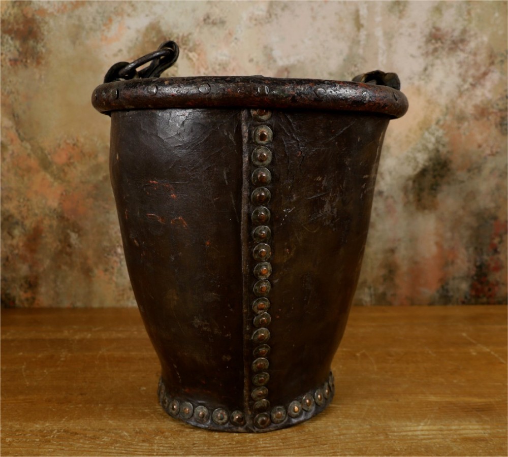 A dark brown Georgian leather fire bucket featuring a heavy strap handle, brass studs around the base, and a painted royal coat of arms with a lion and unicorn. View II