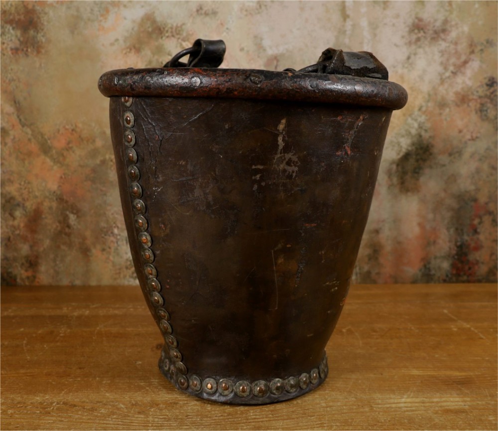 A dark brown Georgian leather fire bucket featuring a heavy strap handle, brass studs around the base, and a painted royal coat of arms with a lion and unicorn. View X