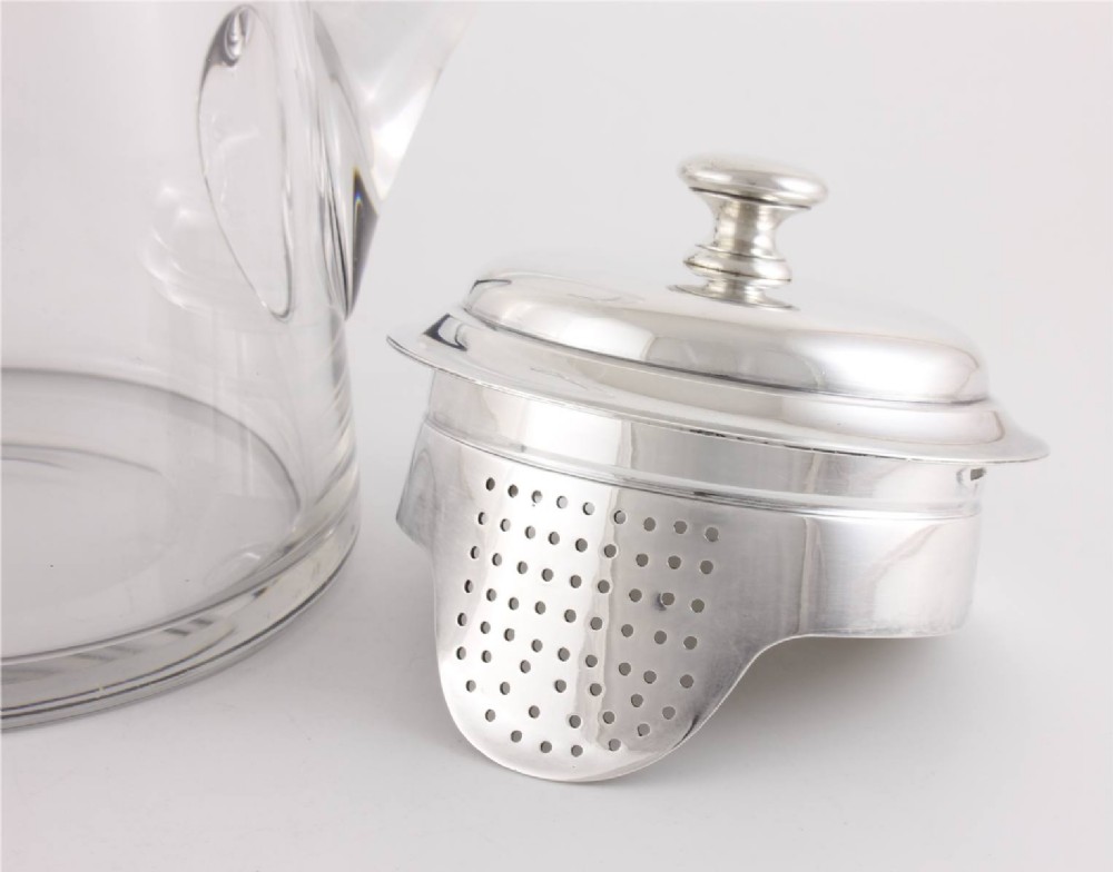 A clear glass water jug with a silver-plated hinged lid, integrated strainer spout, and curved glass handle. View V