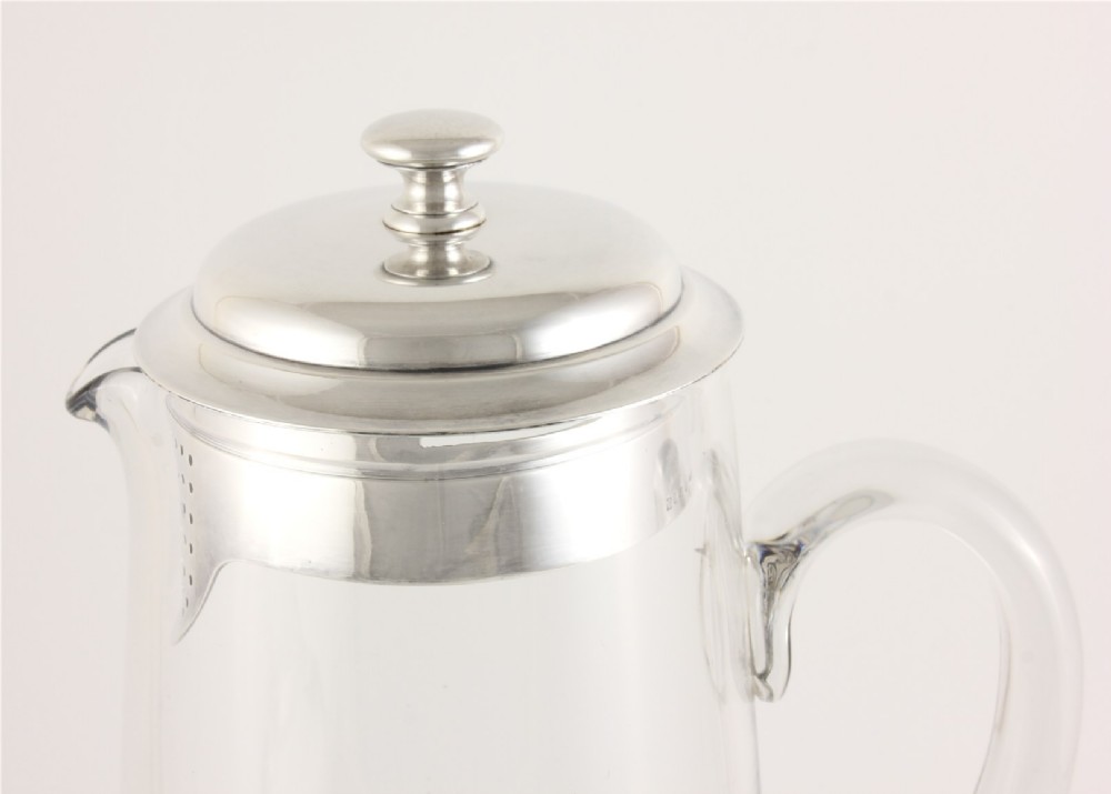 A clear glass water jug with a silver-plated hinged lid, integrated strainer spout, and curved glass handle. View IX