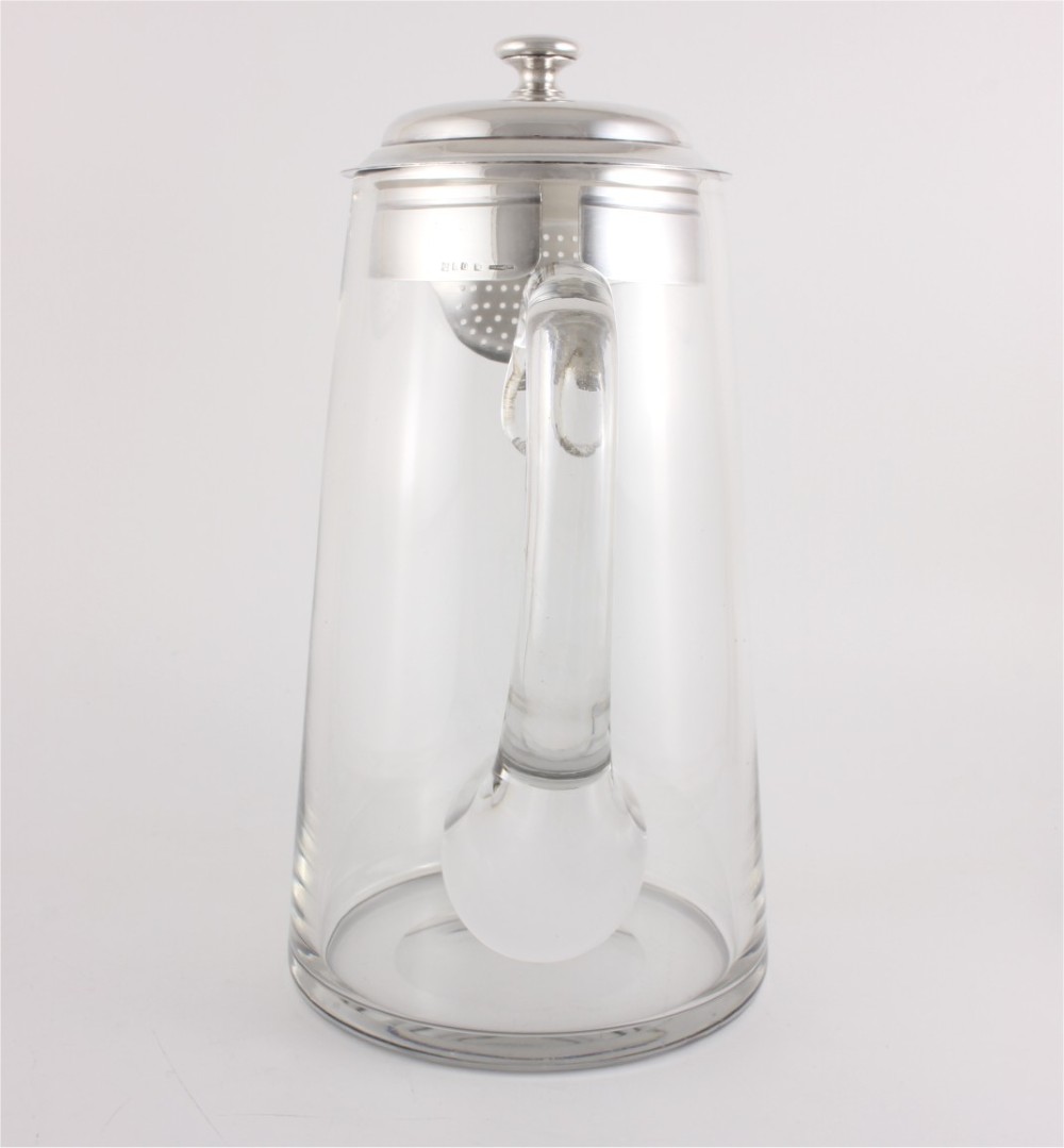 A clear glass water jug with a silver-plated hinged lid, integrated strainer spout, and curved glass handle. View X