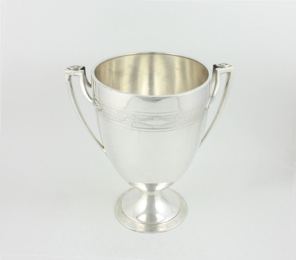 An antique silver-plated urn-shaped champagne cooler featuring twin angular handles, a foliage engraved band, and a stepped pedestal foot.