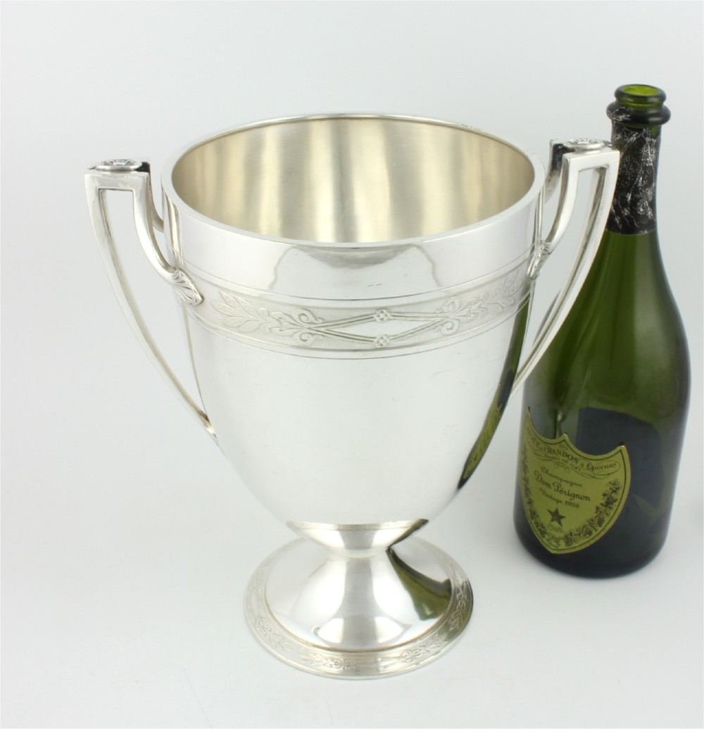 An antique silver-plated urn-shaped champagne cooler featuring twin angular handles, a foliage engraved band, and a stepped pedestal foot. View II