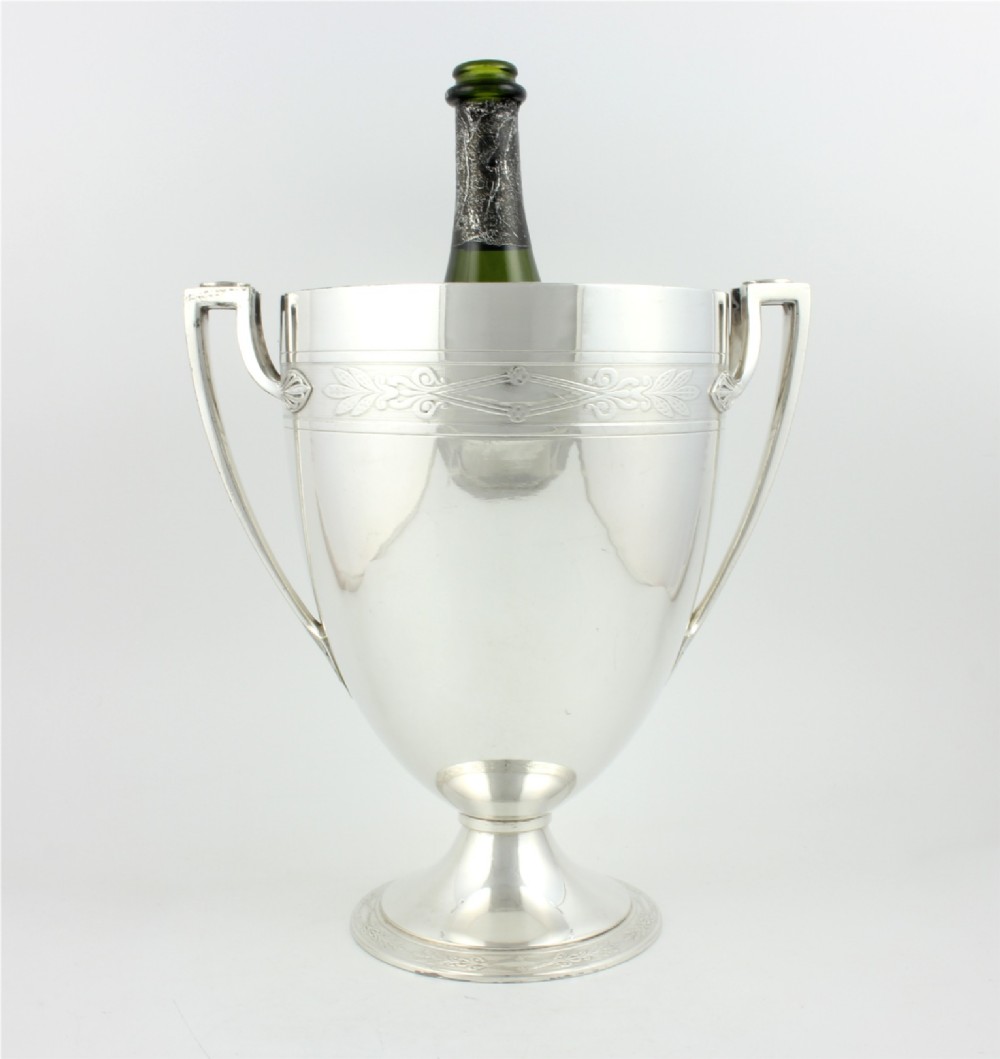An antique silver-plated urn-shaped champagne cooler featuring twin angular handles, a foliage engraved band, and a stepped pedestal foot. View IV