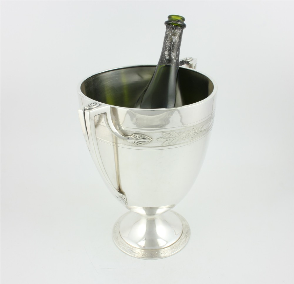An antique silver-plated urn-shaped champagne cooler featuring twin angular handles, a foliage engraved band, and a stepped pedestal foot. View V