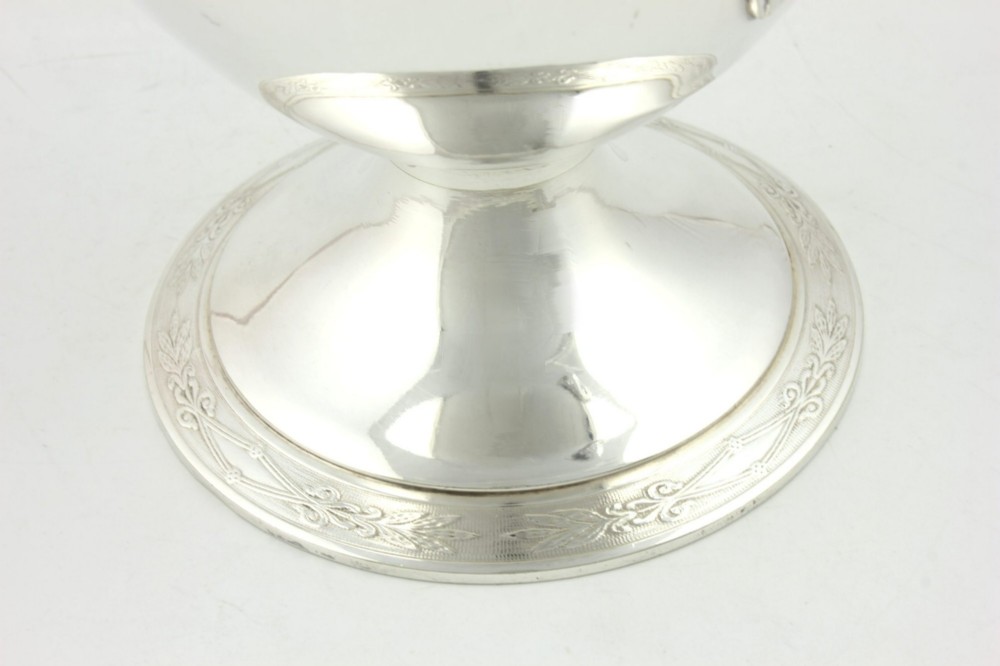 An antique silver-plated urn-shaped champagne cooler featuring twin angular handles, a foliage engraved band, and a stepped pedestal foot. View VII