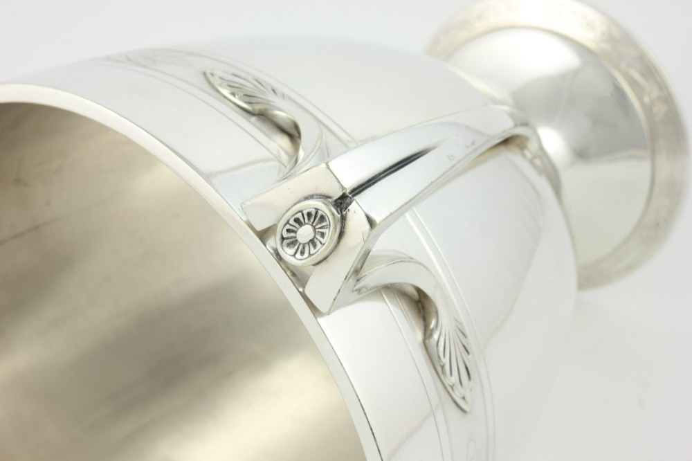 An antique silver-plated urn-shaped champagne cooler featuring twin angular handles, a foliage engraved band, and a stepped pedestal foot. View VIII