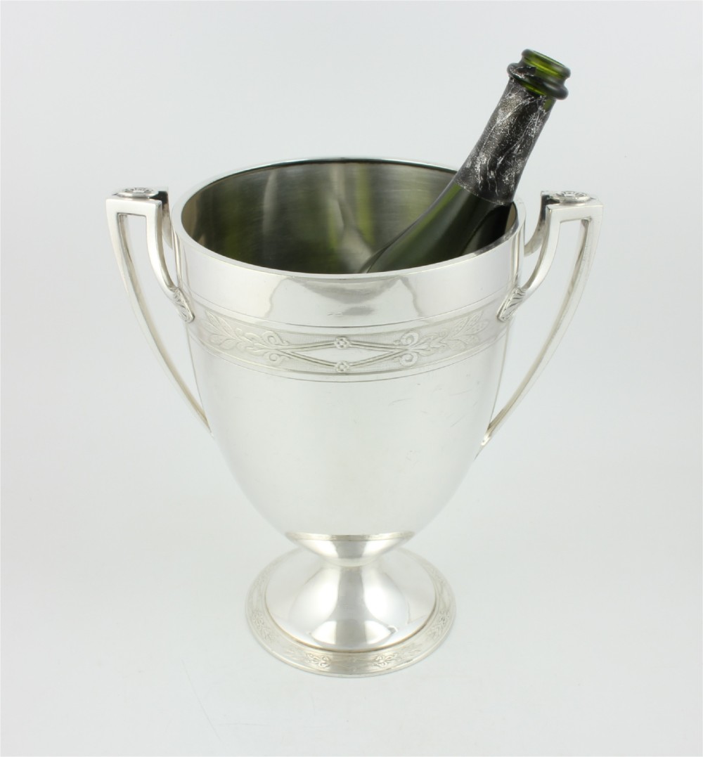 An antique silver-plated urn-shaped champagne cooler featuring twin angular handles, a foliage engraved band, and a stepped pedestal foot. View IX