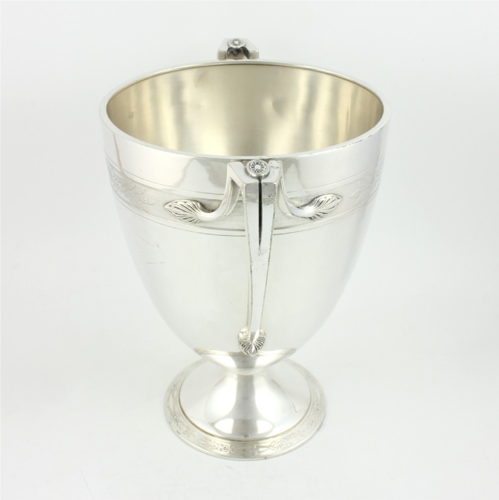 An antique silver-plated urn-shaped champagne cooler featuring twin angular handles, a foliage engraved band, and a stepped pedestal foot. View X