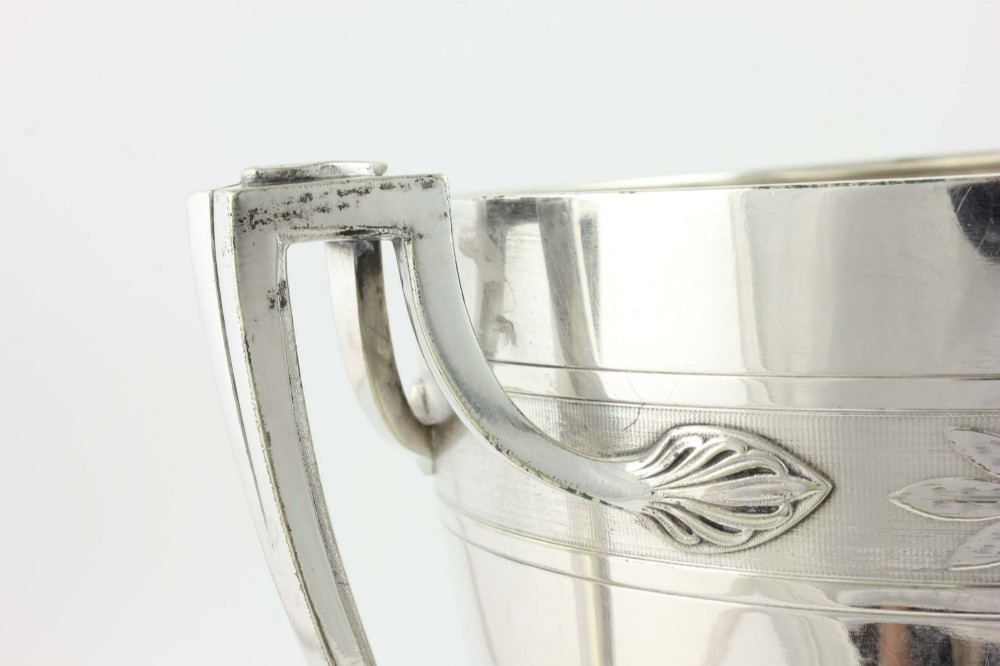 An antique silver-plated urn-shaped champagne cooler featuring twin angular handles, a foliage engraved band, and a stepped pedestal foot. View XIII