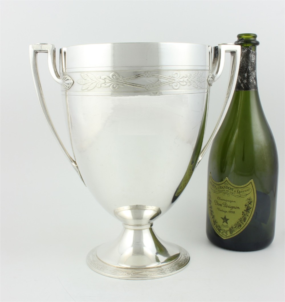 An antique silver-plated urn-shaped champagne cooler featuring twin angular handles, a foliage engraved band, and a stepped pedestal foot. View XIV