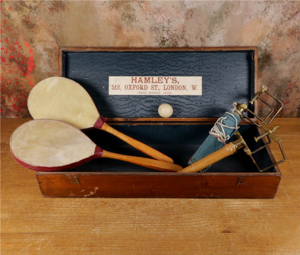An open wooden pine box containing two vellum ping pong rackets, a white ball, and brass net clamps with a folded green net, featuring a Hamleys paper label inside the lid.