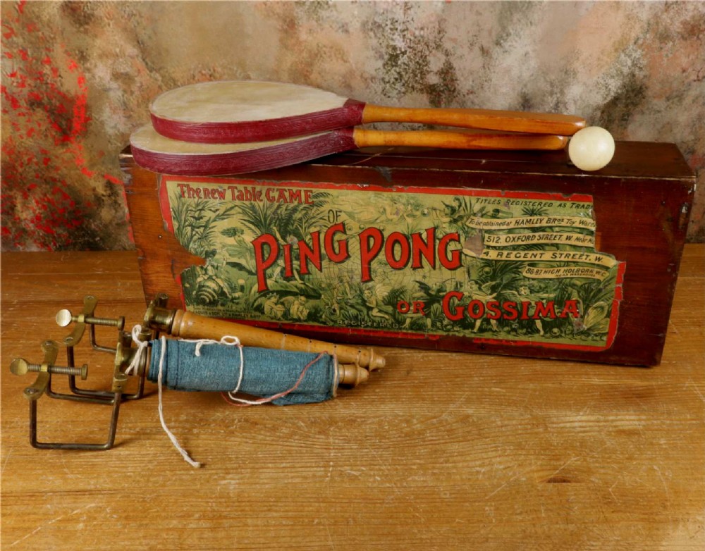 An open wooden pine box containing two vellum ping pong rackets, a white ball, and brass net clamps with a folded green net, featuring a Hamleys paper label inside the lid. View II