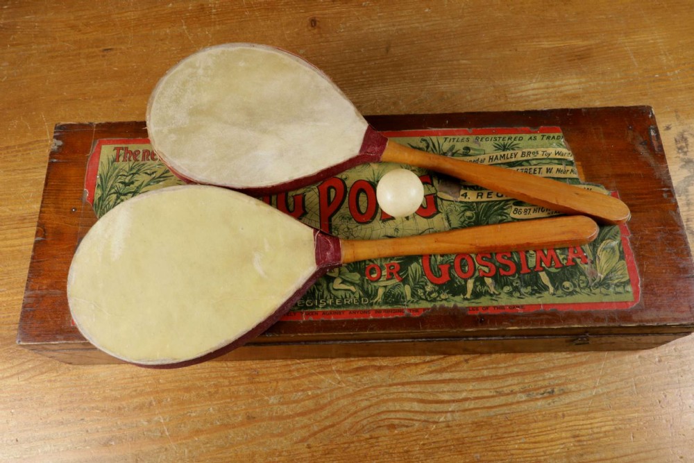 An open wooden pine box containing two vellum ping pong rackets, a white ball, and brass net clamps with a folded green net, featuring a Hamleys paper label inside the lid. View III