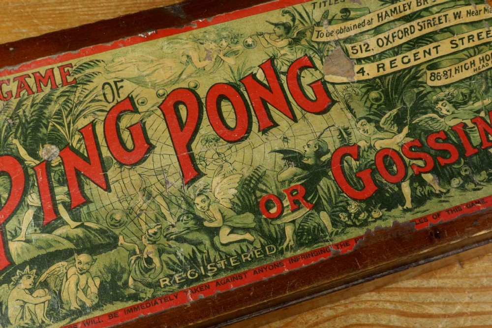 An open wooden pine box containing two vellum ping pong rackets, a white ball, and brass net clamps with a folded green net, featuring a Hamleys paper label inside the lid. View IV