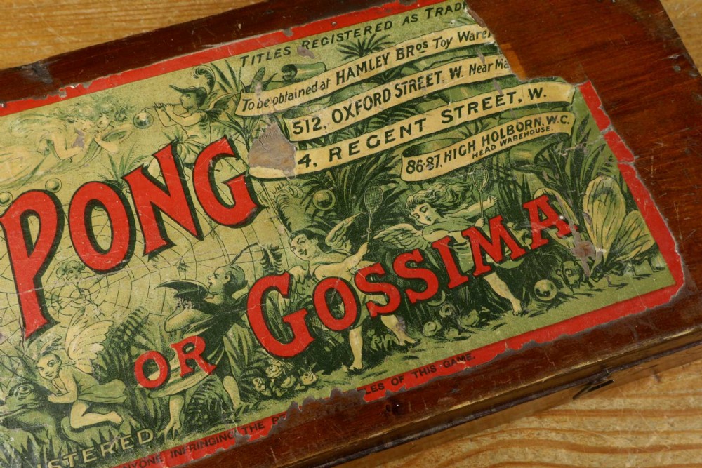 An open wooden pine box containing two vellum ping pong rackets, a white ball, and brass net clamps with a folded green net, featuring a Hamleys paper label inside the lid. View V