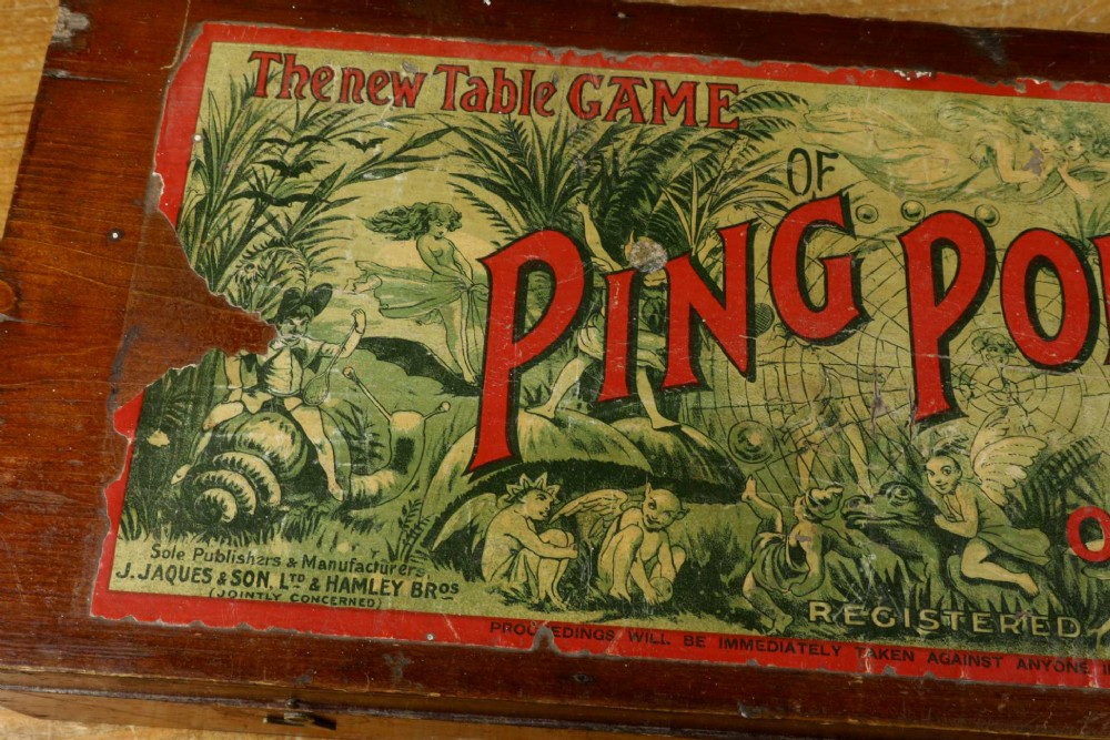 An open wooden pine box containing two vellum ping pong rackets, a white ball, and brass net clamps with a folded green net, featuring a Hamleys paper label inside the lid. View VI