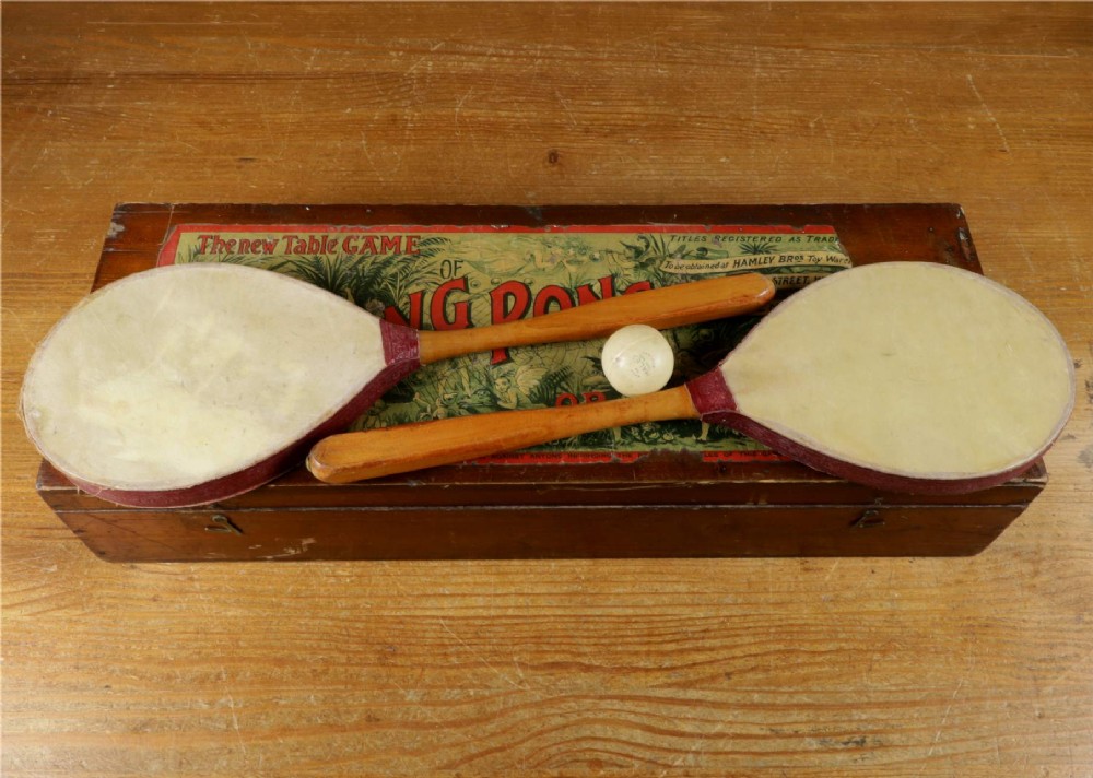 An open wooden pine box containing two vellum ping pong rackets, a white ball, and brass net clamps with a folded green net, featuring a Hamleys paper label inside the lid. View VII