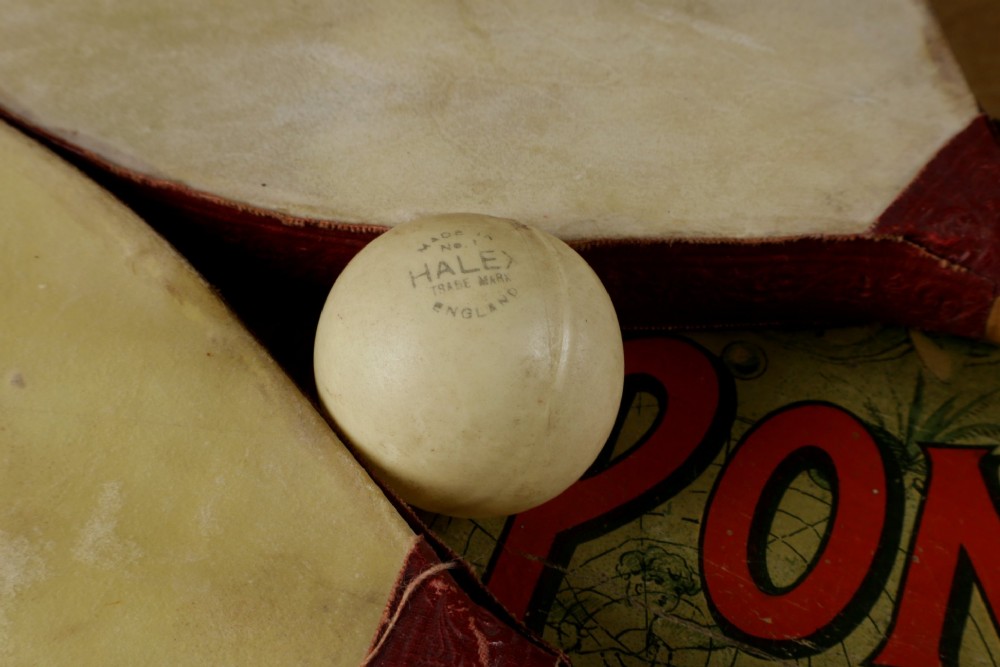 An open wooden pine box containing two vellum ping pong rackets, a white ball, and brass net clamps with a folded green net, featuring a Hamleys paper label inside the lid. View X