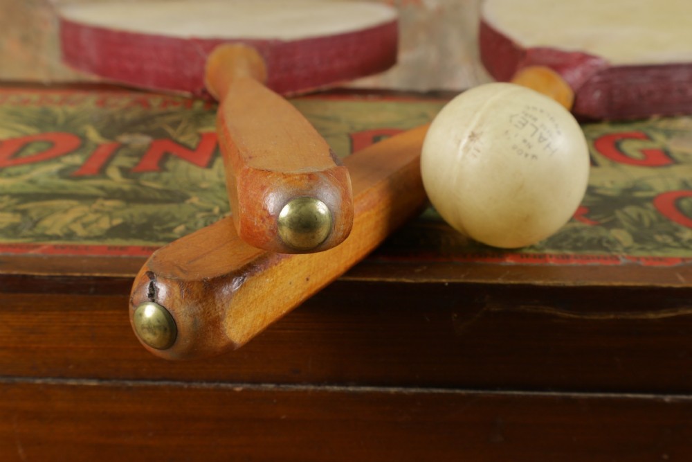 An open wooden pine box containing two vellum ping pong rackets, a white ball, and brass net clamps with a folded green net, featuring a Hamleys paper label inside the lid. View XI