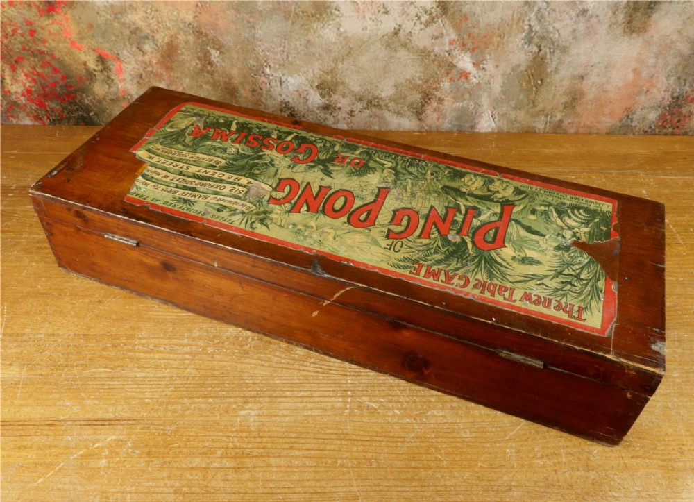 An open wooden pine box containing two vellum ping pong rackets, a white ball, and brass net clamps with a folded green net, featuring a Hamleys paper label inside the lid. View XIII