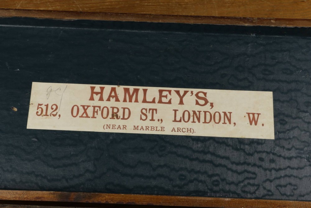 An open wooden pine box containing two vellum ping pong rackets, a white ball, and brass net clamps with a folded green net, featuring a Hamleys paper label inside the lid. View XIV