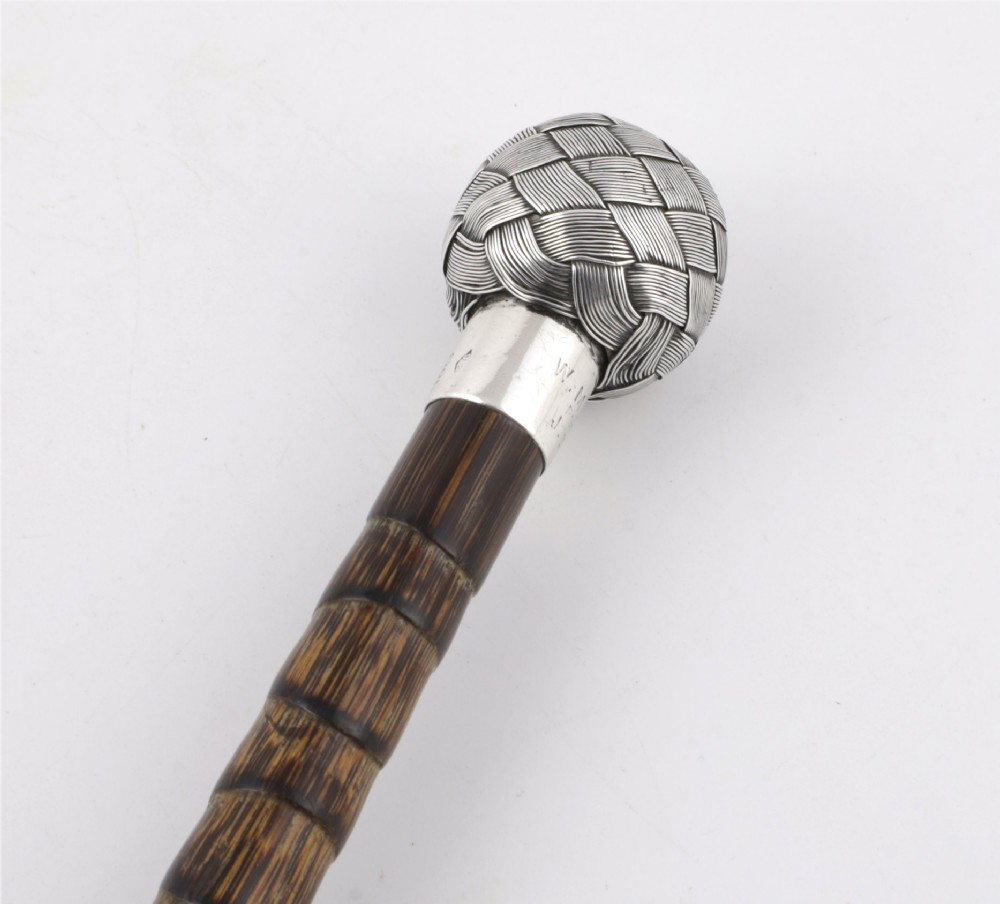 A close-up of an antique walking stick featuring a woven silver wire knot handle mounted on a dark bamboo shaft with a silver collar.