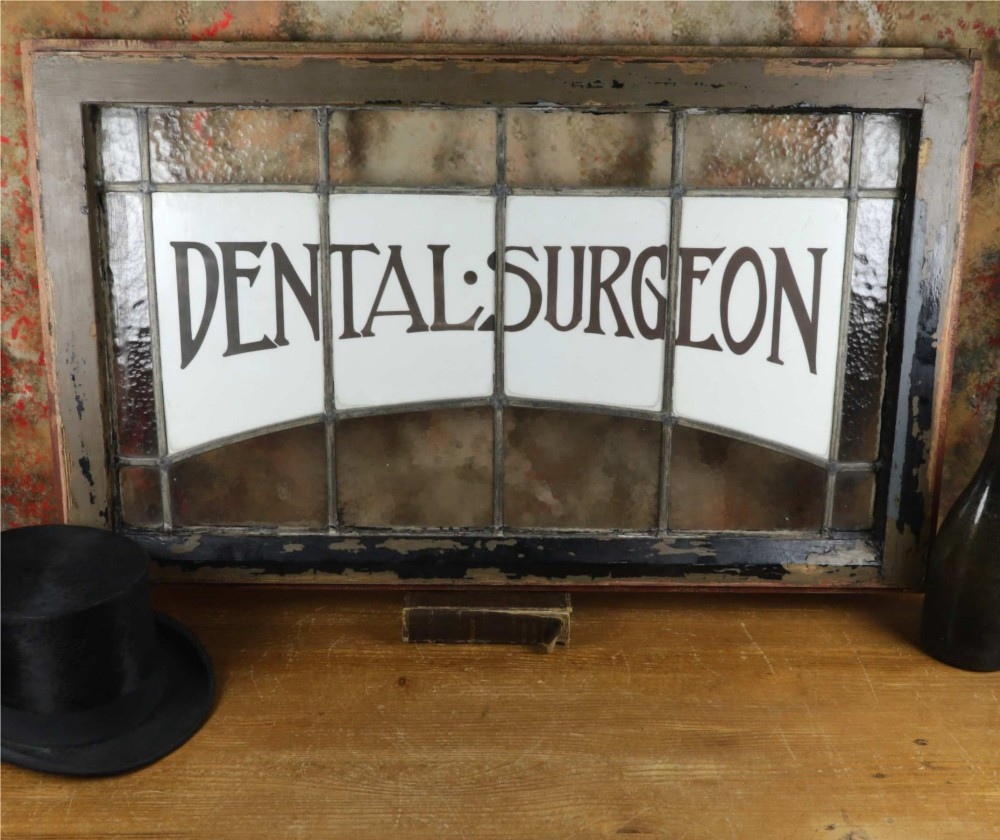 An Edwardian leaded glass window panel set in a dark wooden frame with white glass panes featuring black lettering that reads DENTAL SURGEON.