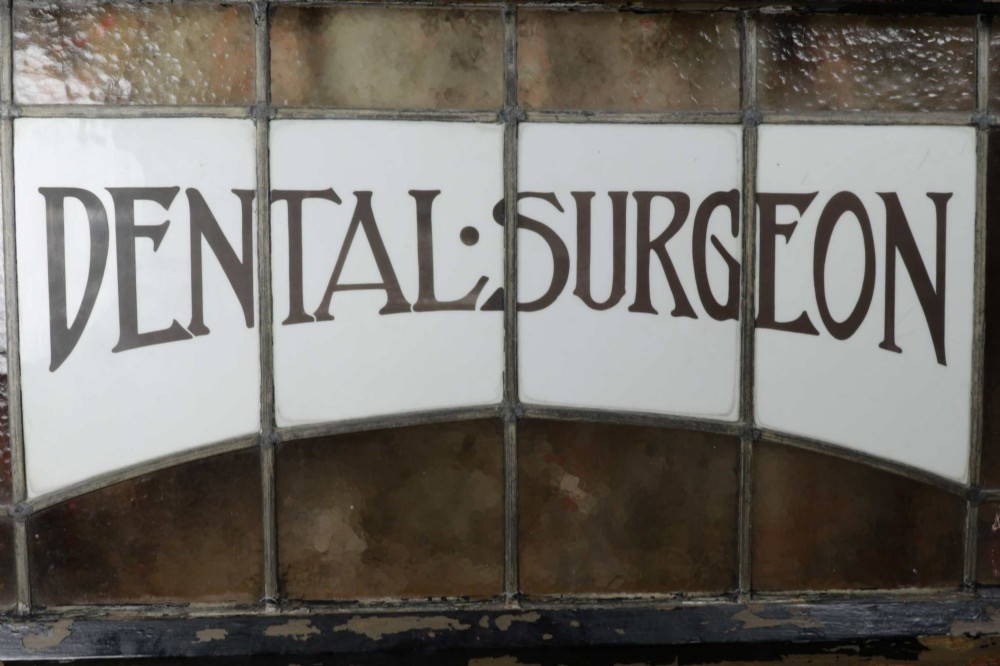 An Edwardian leaded glass window panel set in a dark wooden frame with white glass panes featuring black lettering that reads DENTAL SURGEON. View II