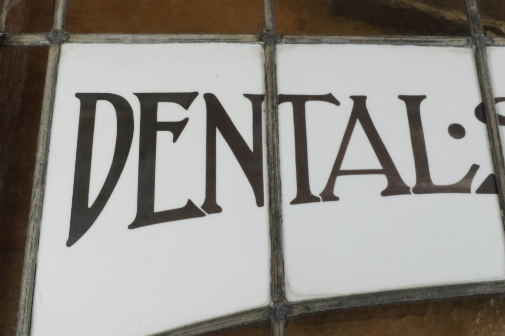 An Edwardian leaded glass window panel set in a dark wooden frame with white glass panes featuring black lettering that reads DENTAL SURGEON. View V