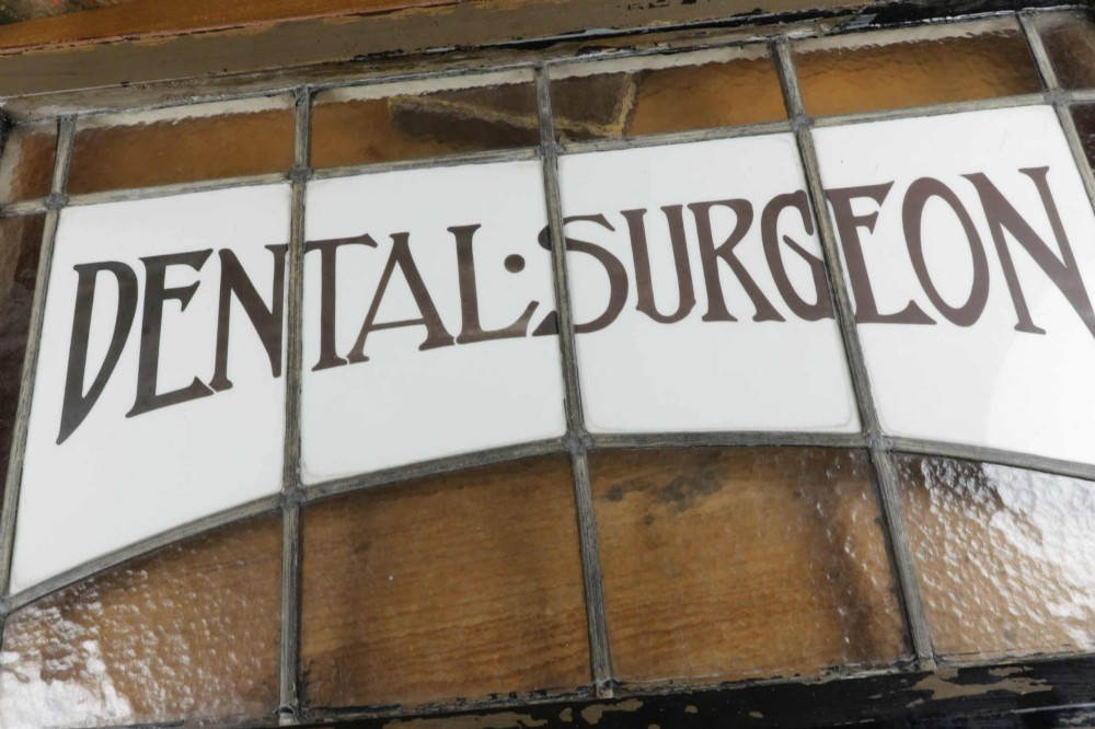 An Edwardian leaded glass window panel set in a dark wooden frame with white glass panes featuring black lettering that reads DENTAL SURGEON. View VII