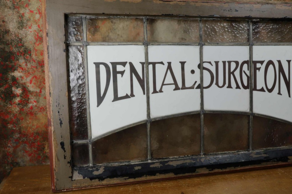 An Edwardian leaded glass window panel set in a dark wooden frame with white glass panes featuring black lettering that reads DENTAL SURGEON. View XII