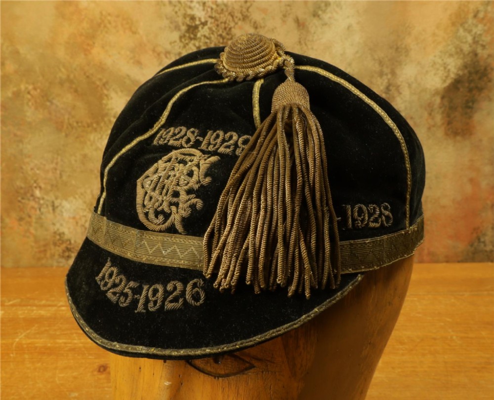 A 1920s black velvet sports cap featuring metallic braid piping, embroidered dates, monogram crest, and a prominent hanging tassel.