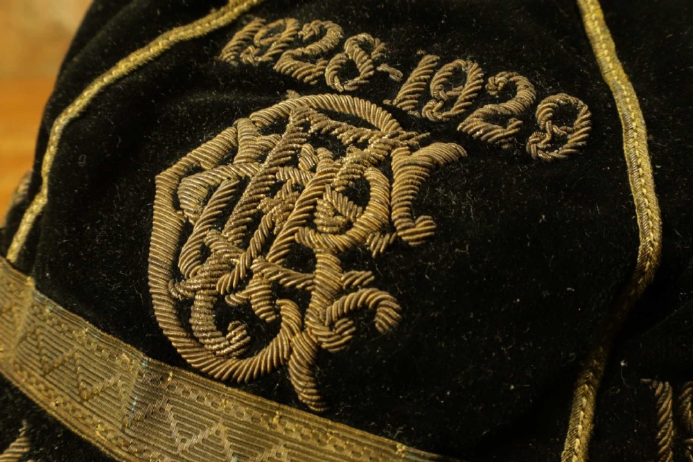 A 1920s black velvet sports cap featuring metallic braid piping, embroidered dates, monogram crest, and a prominent hanging tassel. View IV
