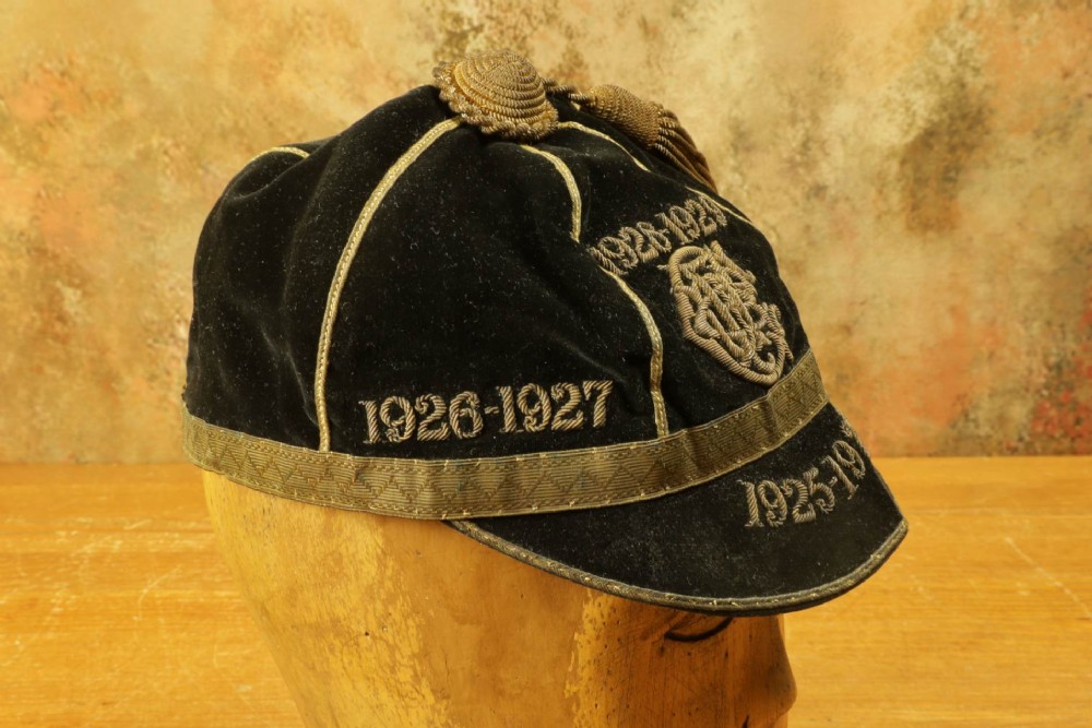 A 1920s black velvet sports cap featuring metallic braid piping, embroidered dates, monogram crest, and a prominent hanging tassel. View V