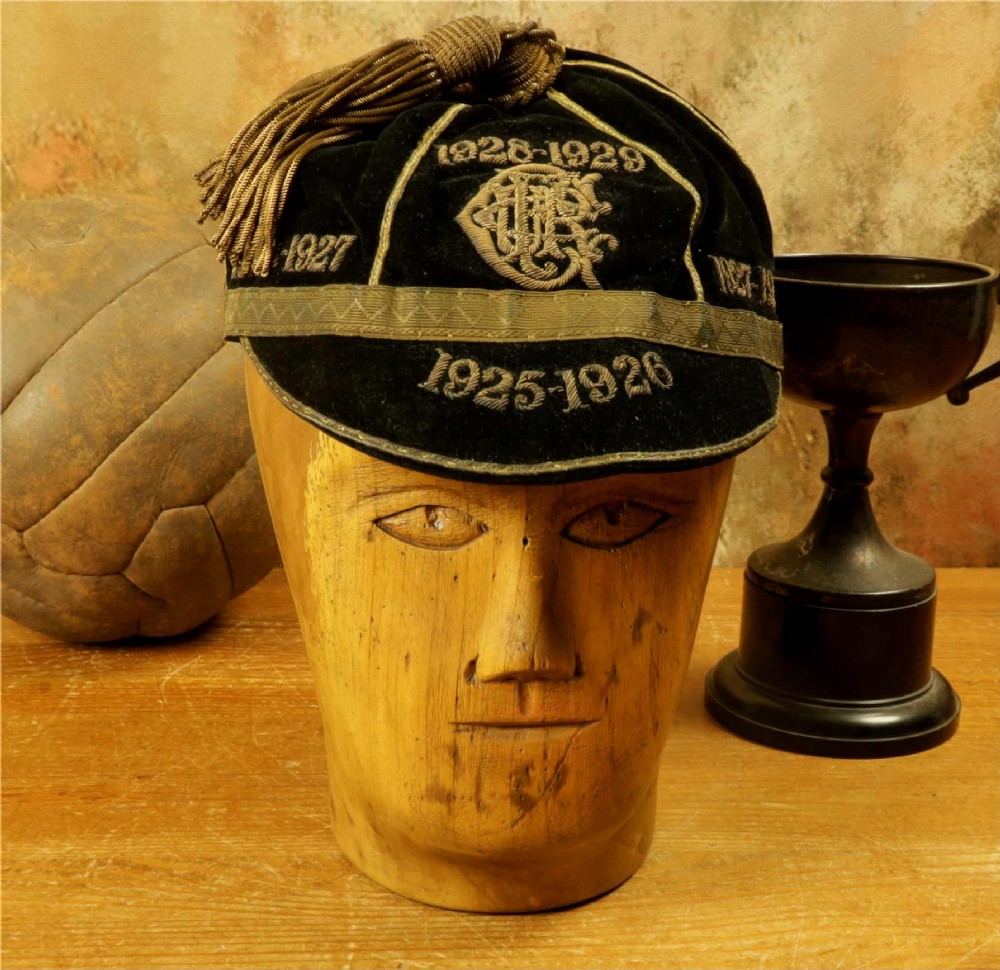 A 1920s black velvet sports cap featuring metallic braid piping, embroidered dates, monogram crest, and a prominent hanging tassel. View VI