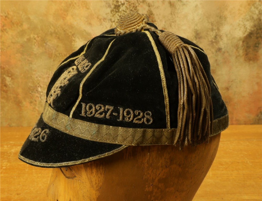 A 1920s black velvet sports cap featuring metallic braid piping, embroidered dates, monogram crest, and a prominent hanging tassel. View VII