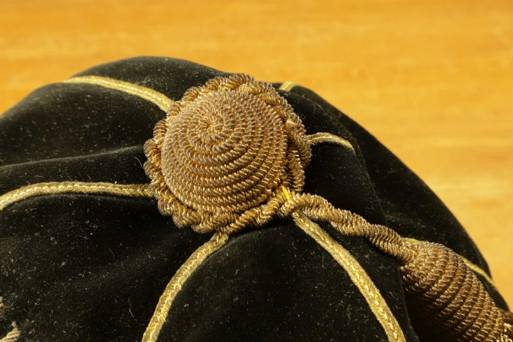 A 1920s black velvet sports cap featuring metallic braid piping, embroidered dates, monogram crest, and a prominent hanging tassel. View VIII