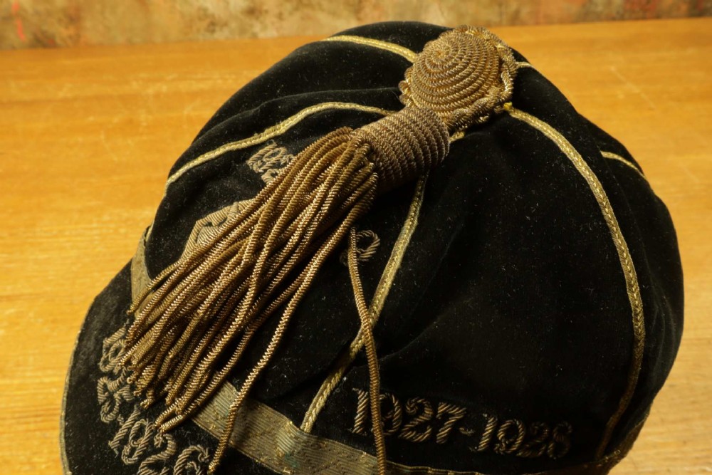 A 1920s black velvet sports cap featuring metallic braid piping, embroidered dates, monogram crest, and a prominent hanging tassel. View IX