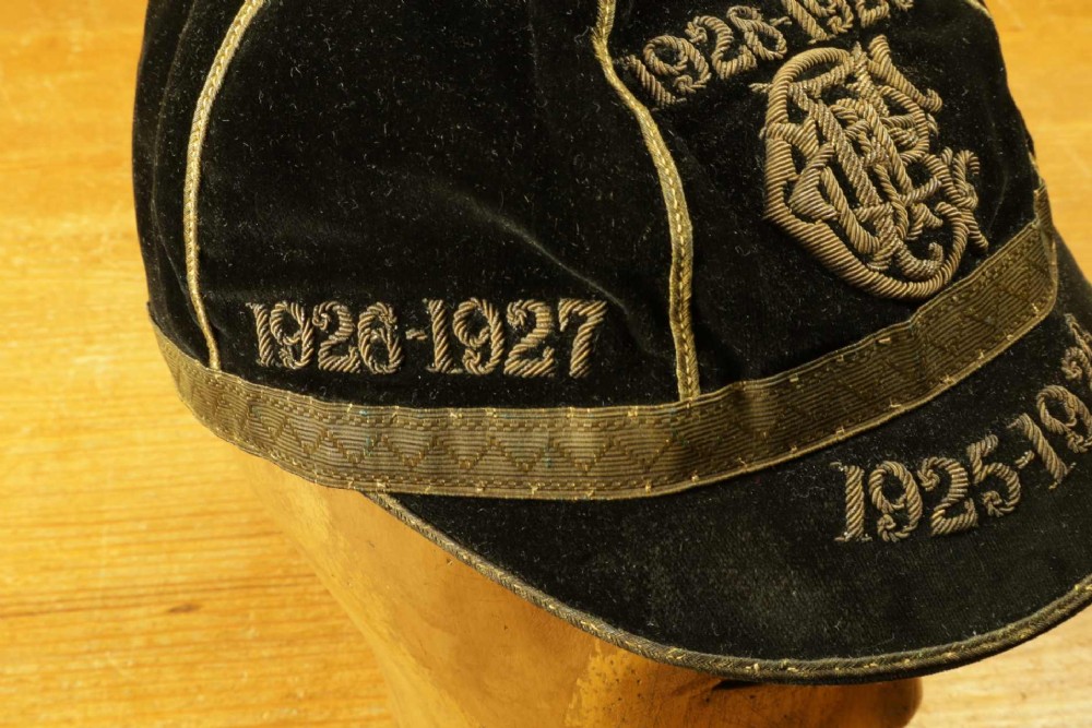 A 1920s black velvet sports cap featuring metallic braid piping, embroidered dates, monogram crest, and a prominent hanging tassel. View X