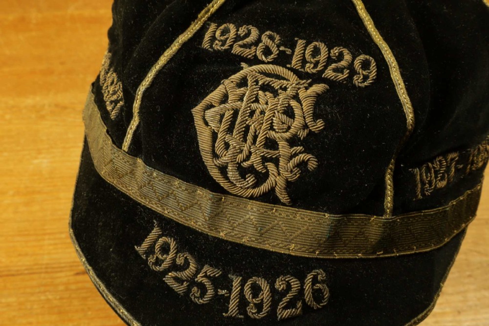 A 1920s black velvet sports cap featuring metallic braid piping, embroidered dates, monogram crest, and a prominent hanging tassel. View XIV