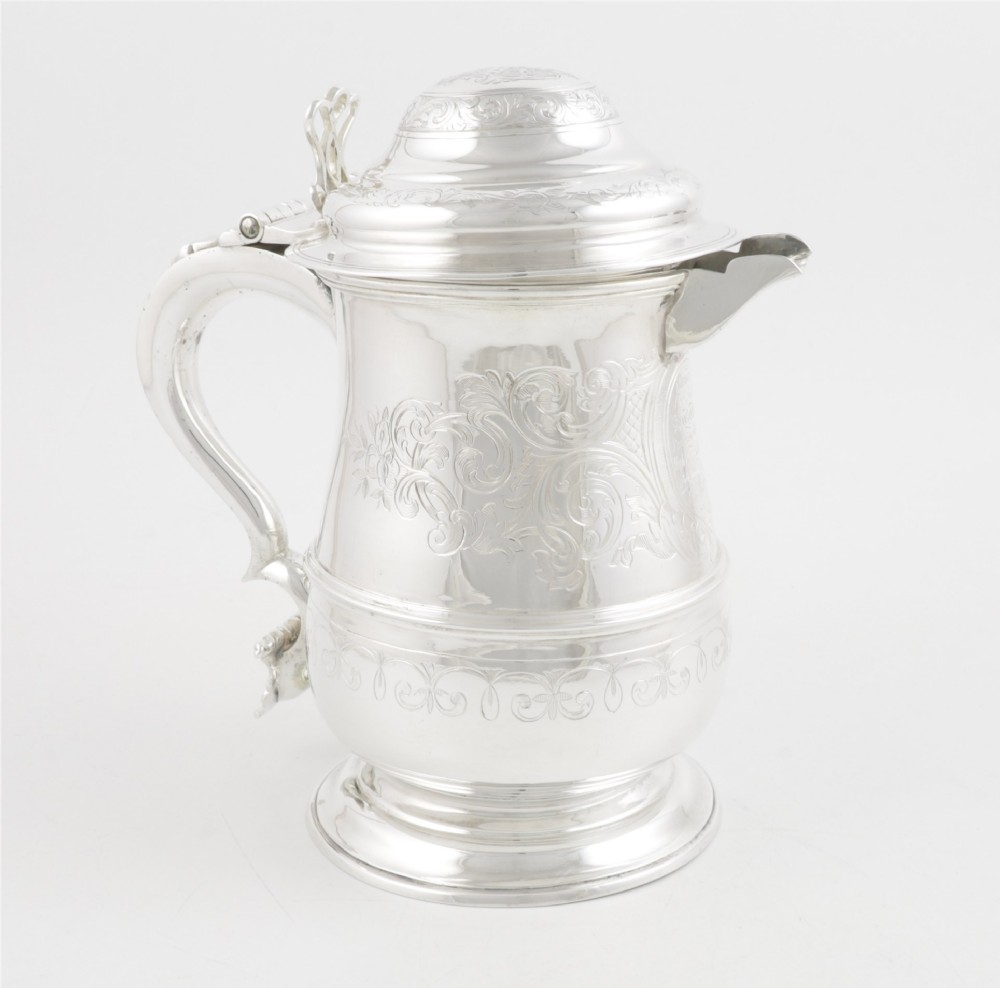 An engraved silver lidded jug featuring a hinged cap, scrolling handle, and foliate engraving across the body.
